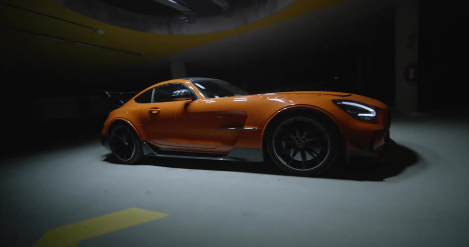 An FPV drone shot zooming around an orange luxury sports car parked under a spotlight inside a dark parking garage