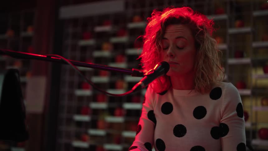 A woman sings into a microphone while playing the keyboard under a red light indoors