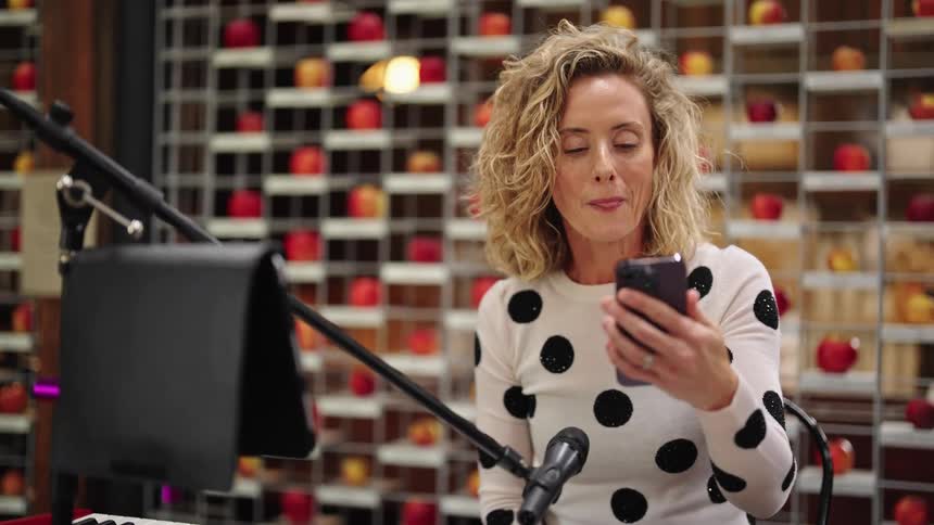 A woman is sitting behind a microphone as she holds her phone in her hand and waves to someone as she speaks