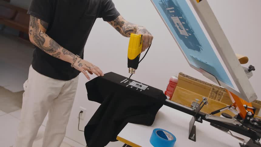 A man is standing over a silk screened tshirt drying it with a dryer