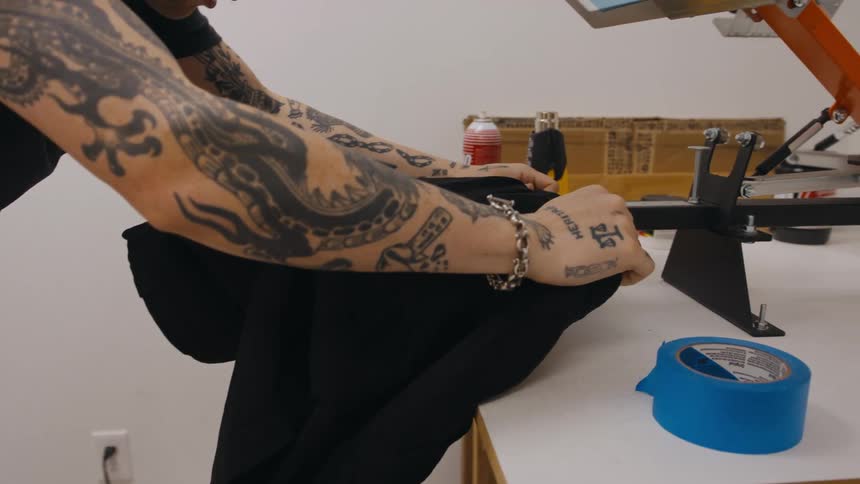 Young tattooed man places a black tshirt on the table of a screen printing press