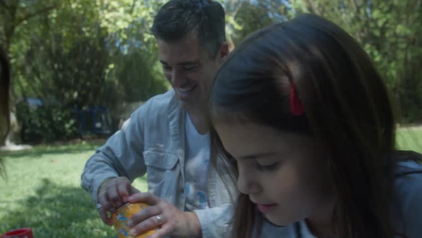 A man and a woman are sitting down in the grass with their daughter painting a big picture with a roller and a brush