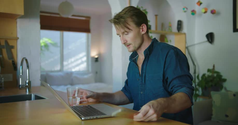 A man types on a laptop and sips on water in a kitchen