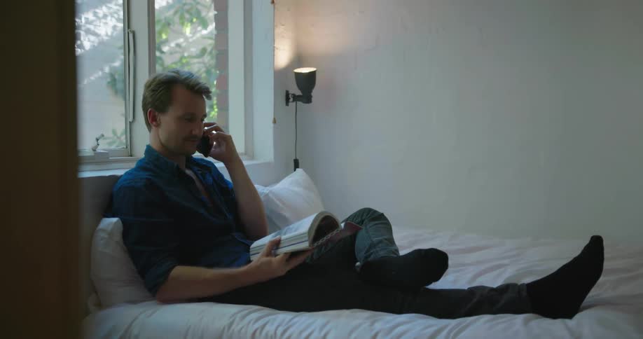 Man sitting on bed holding book and talking on cell phone during the day
