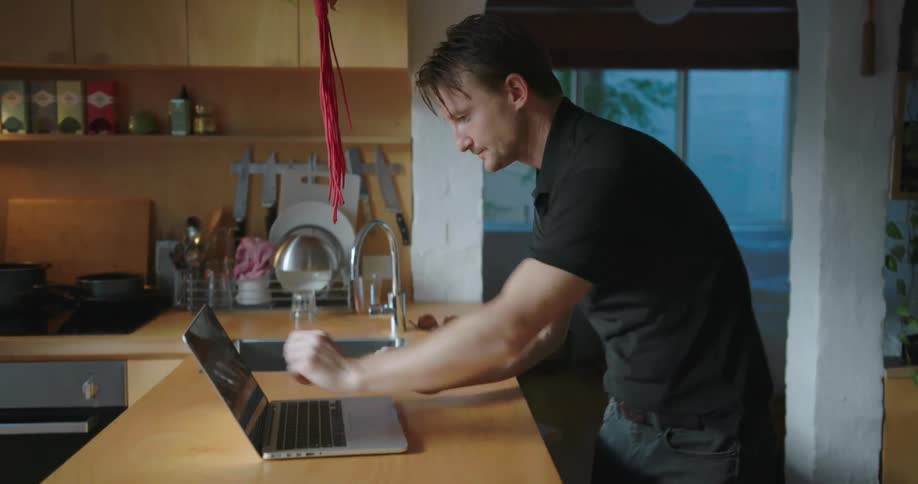 A man puts on a shirt and walks towards a laptop and opens it