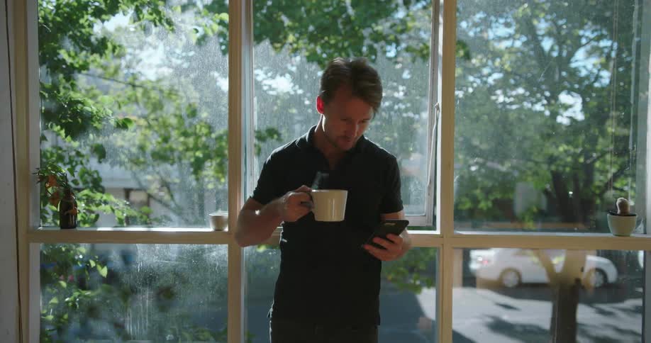 A man sips coffee and looks at his phone while standing next to a window