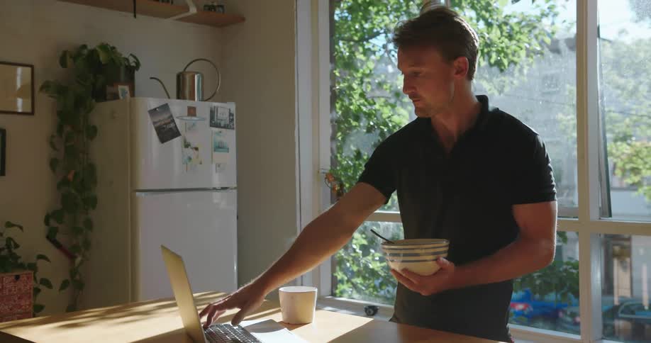 Man stands at a kitchen counter drinking out of a mug, eating and typing on a laptop