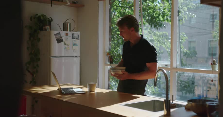 A man eats from a bowl while looking at a laptop on the kitchen counter
