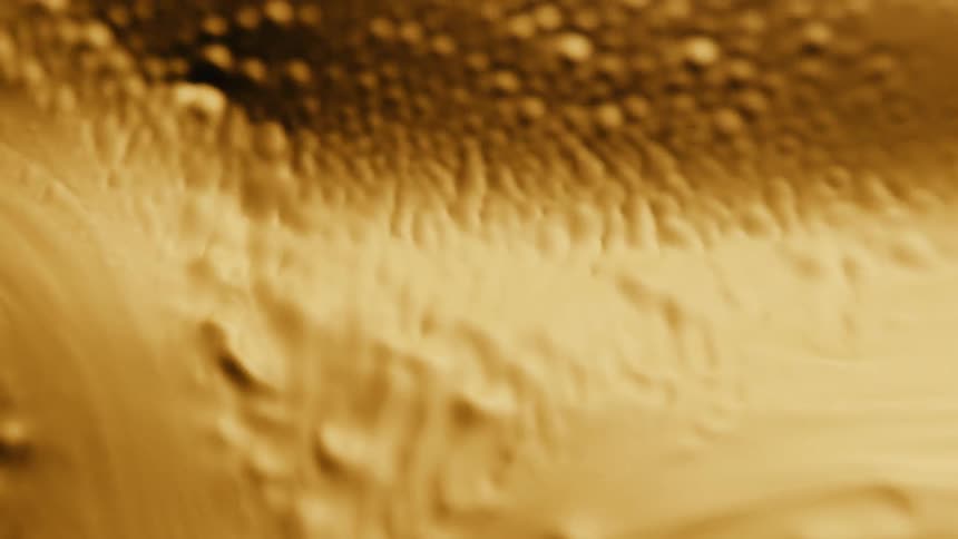 Light shines on a gold organic liquid substance as bubbles and holes emerge as it ripples and moves and then melts down