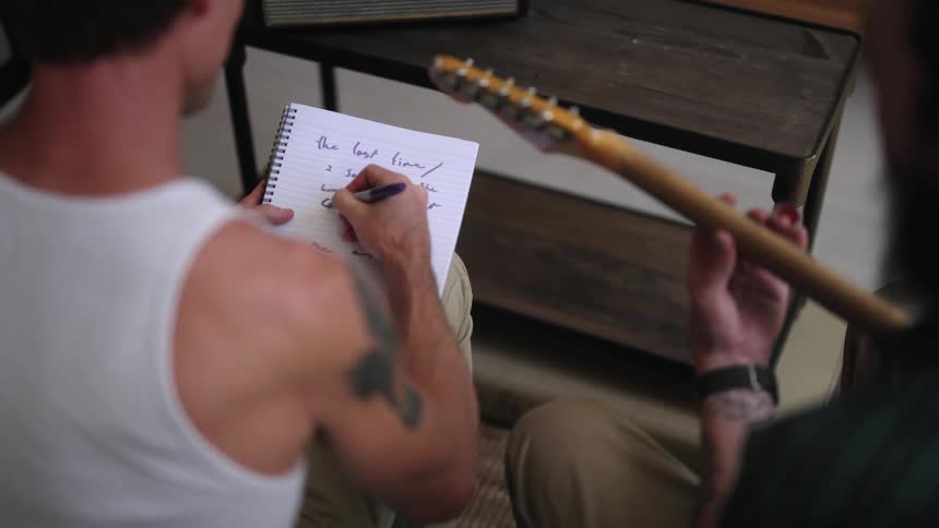 A man sits and writes in a notebook as the man next to him plays the guitar