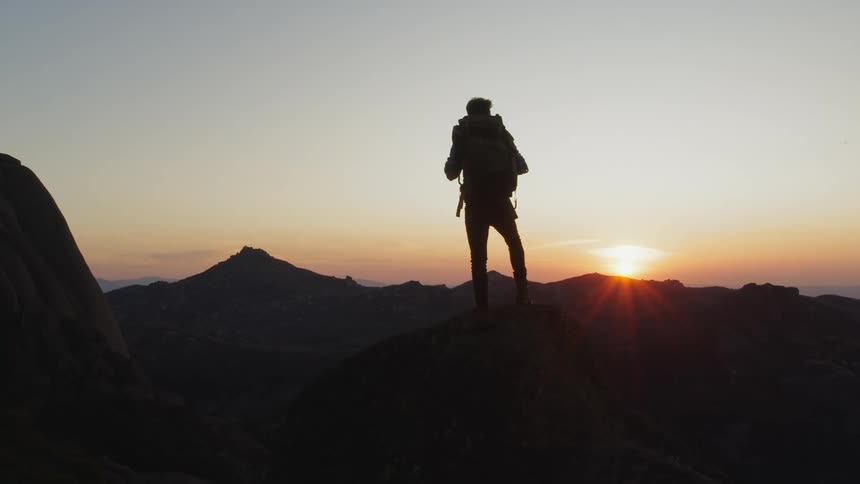 Man silhouetted by the rising sun stands on peak staring to the sun