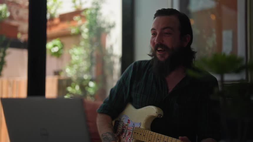 A man on laptop video call holds electric guitar