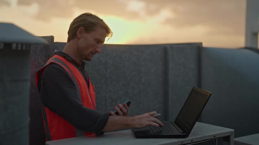 A construction worker using a laptop and smartphone