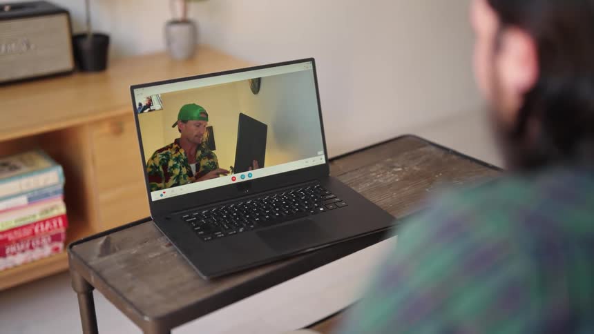 Man with beard and plaid shirt makes video call on laptop with man wearing green cap and shirt in room