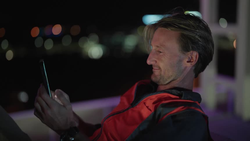 Middle aged blonde man wearing black and red jacket sits in chair texting on smartphone outdoors