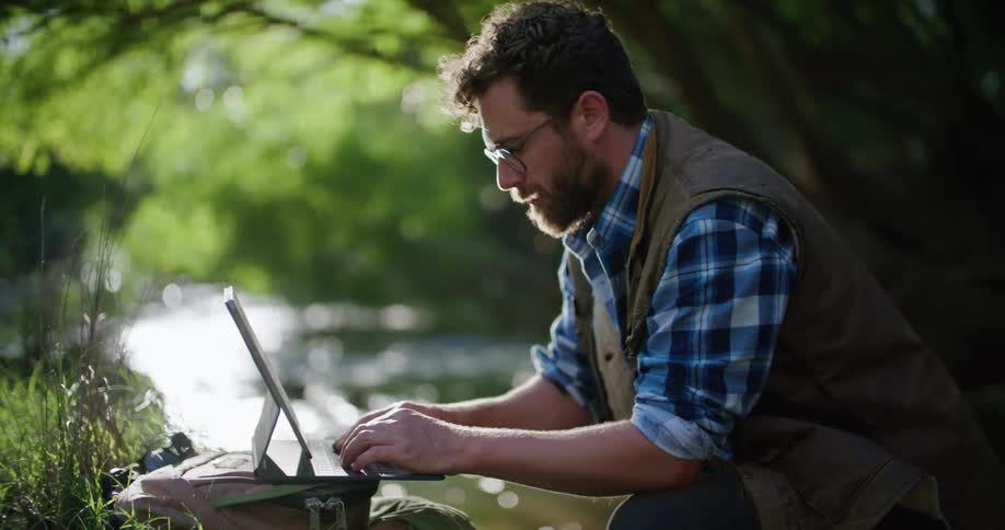 A man is kneeling in the woods near a stream as he types on his laptop computer