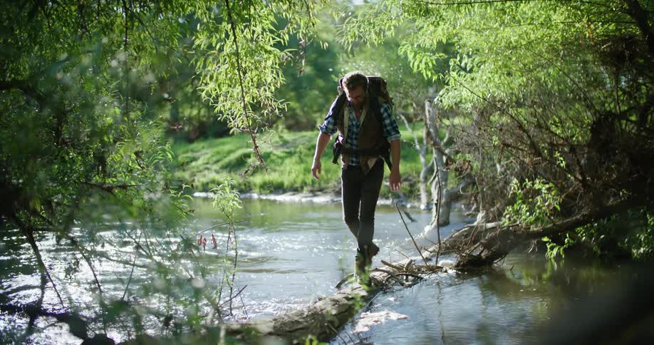 A man is hiking in the woods and crosses over a river by using a fallen tree trunk as a bridge
