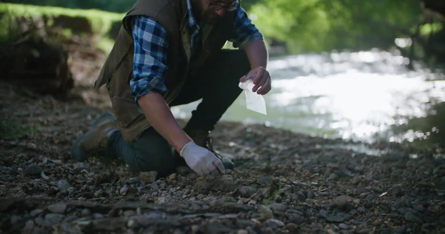Man wearing glasses and a vest kneels near a stream and collects rocks in a small bag