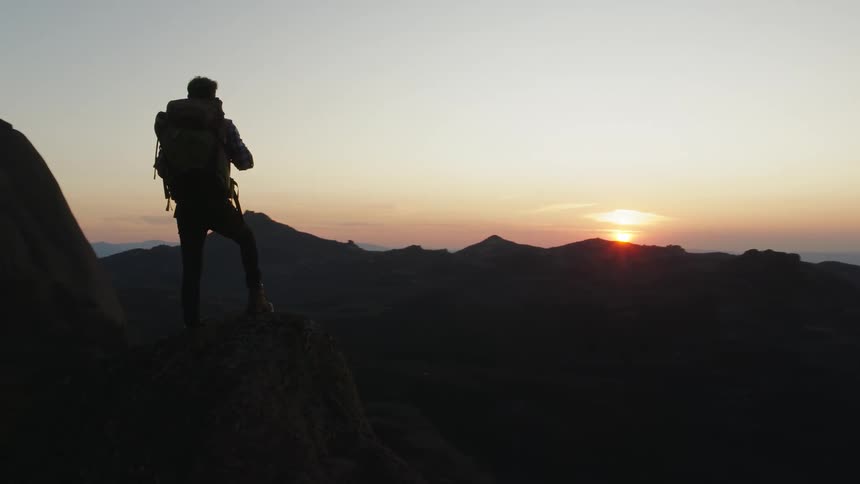 A man hiking in a mountain range stands and looks at the sun rising