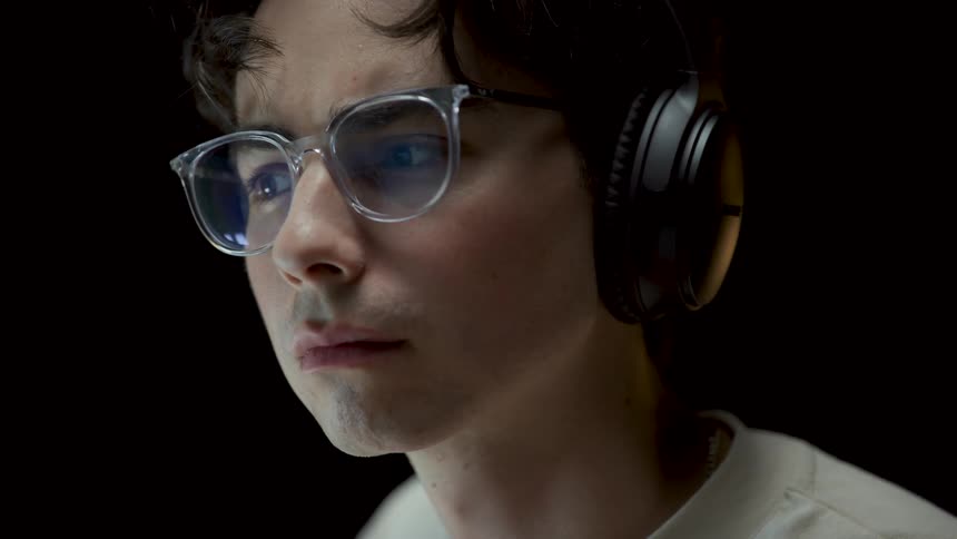 Light shines on the face of a boy wearing glasses and headphones sitting in a dark room