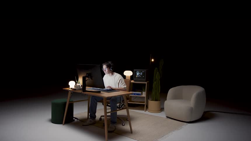 A man wearing headphones is sitting at a desk typing on a computer keyboard and there is living room furniture in a wide open room