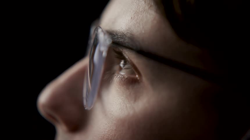 Close up of the face of a man wearing glasses looking up in front of a black backdrop