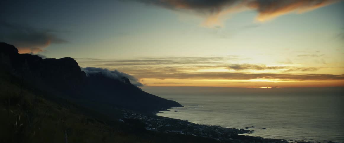 Fog rolls over rocks along the coast as waves roll over the beach and clouds move across the sky as the sun sets on the horizon