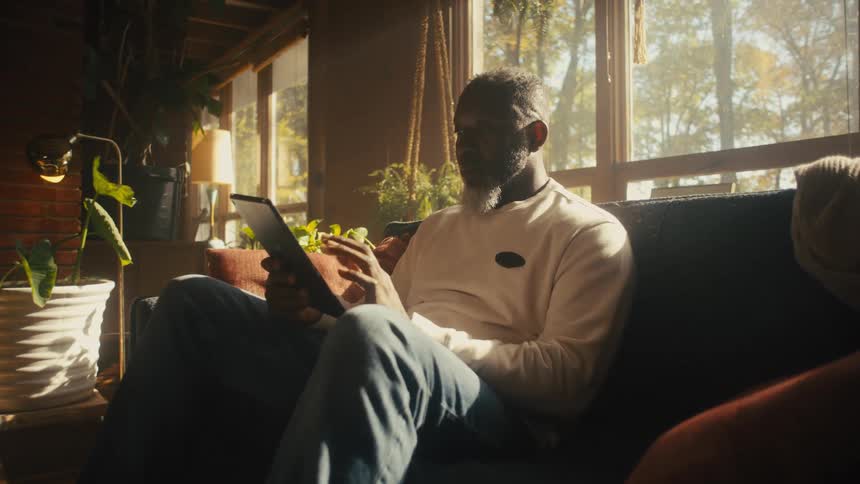 A man sitting on a living room sofa while scrolling on a tablet