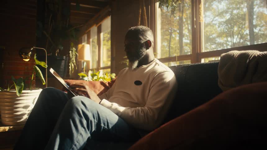 Older man sits on couch in living room using tablet on sunny day