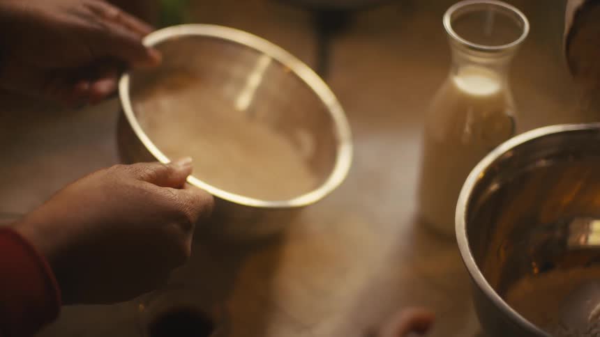 A person measuring out a cupful of flour and pouring it into a metal mixing bowl