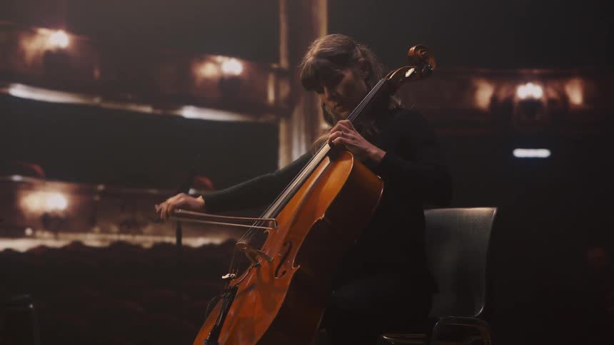 A woman is seated on stage playing cello in a lit theatre