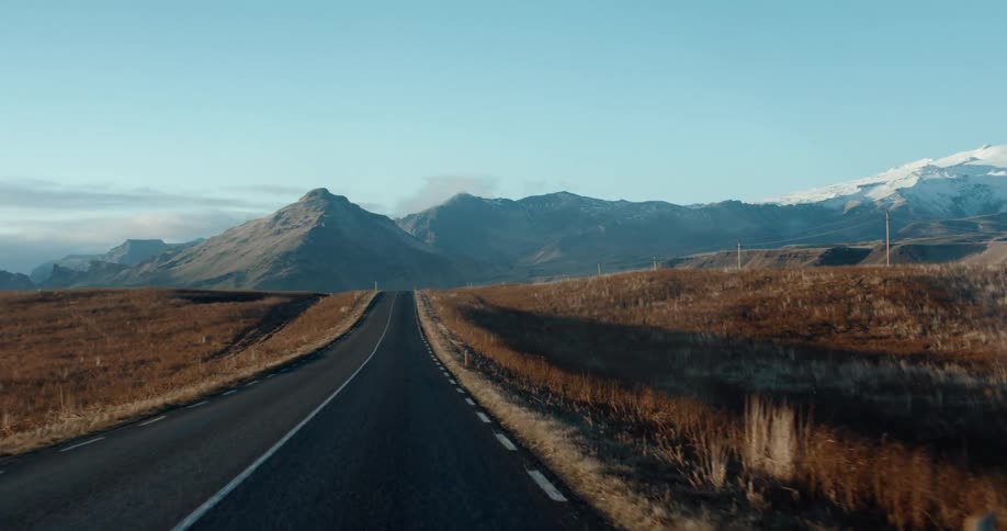 Driving towards the mountains on a straight paved road with dry grass on the roadside