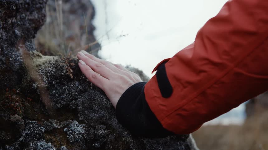 Woman wearing orange coat touches large rock on the coast