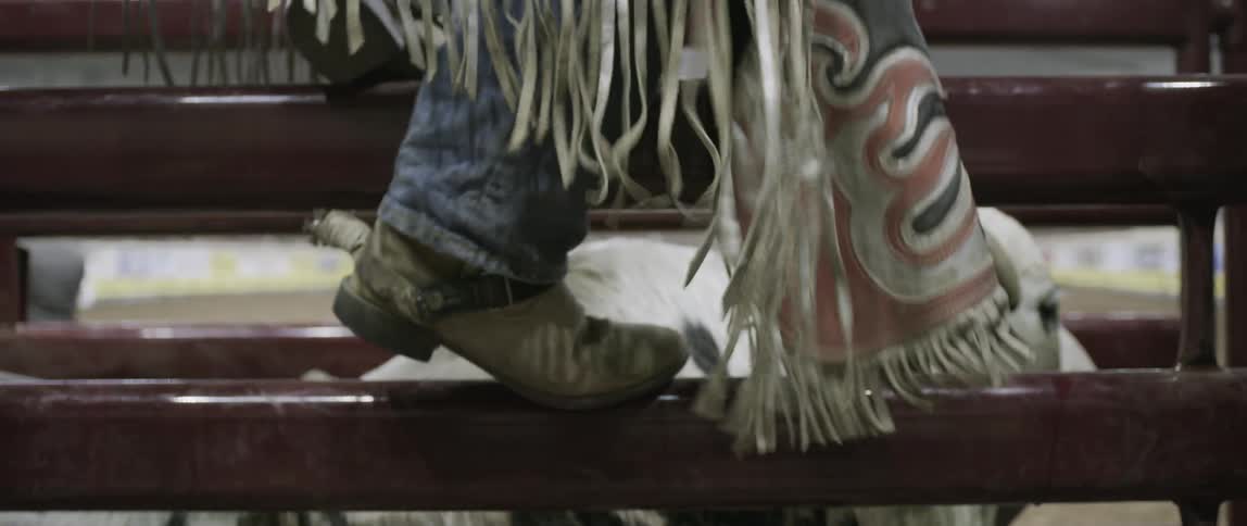 The fringe on a pair of chaps swaying as a man shakes his leg while standing on the side of a rodeo bull pen