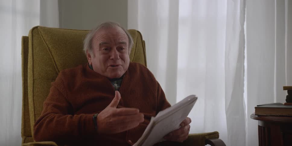 A man is sitting in a chair holding a notebook as he speaks to someone across the room