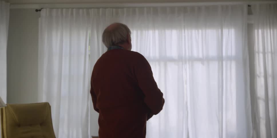 An elderly man walking towards curtained windows at home