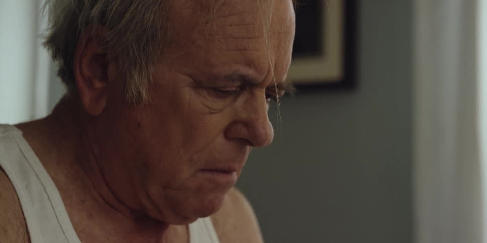 A man is sitting down on the edge of his bed looking at paperwork and he stops to take off his glasses and turn his head