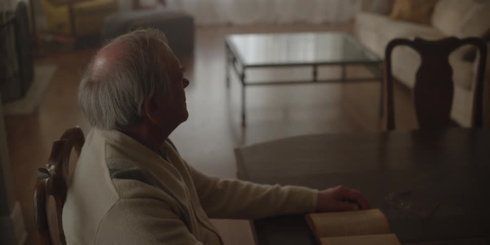 Man sits at dining room table alone with book and stares across area