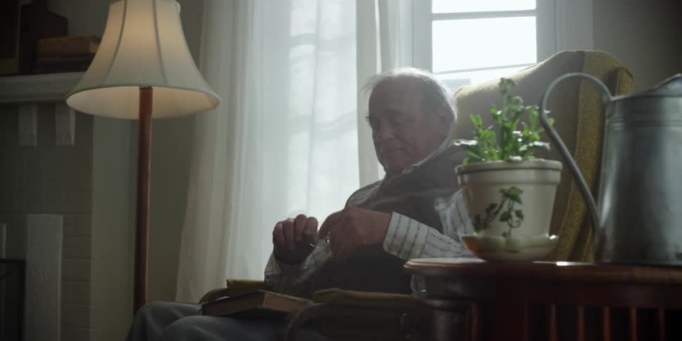 An elderly man putting on glasses to read a book while sitting in a living room chair