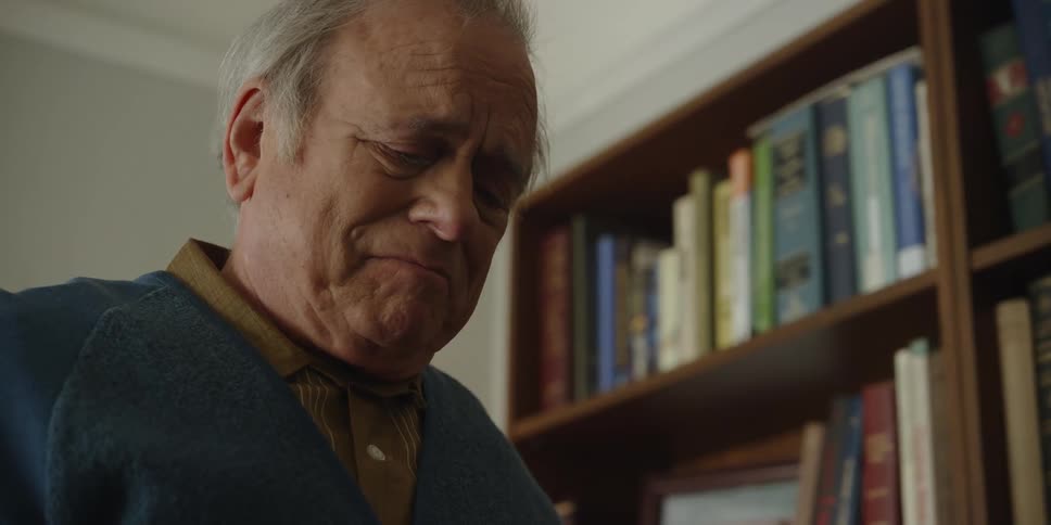 A man is standing near a bookcase reflecting on something that is making him sad and he walks away
