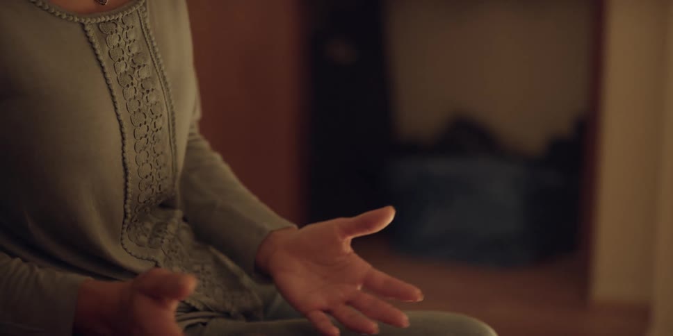 A woman speaks with her hands animatedly while sitting on the side of a bed