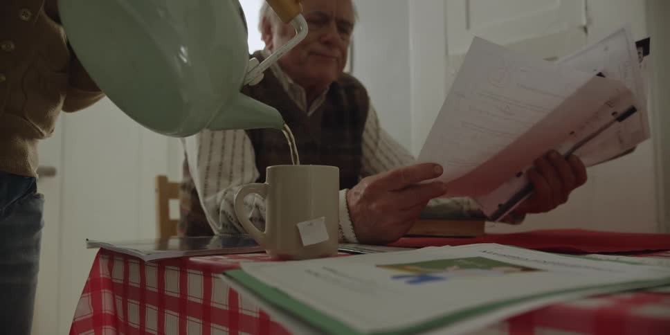 An elderly man shakes his head as he looks through papers while a woman pours him some tea