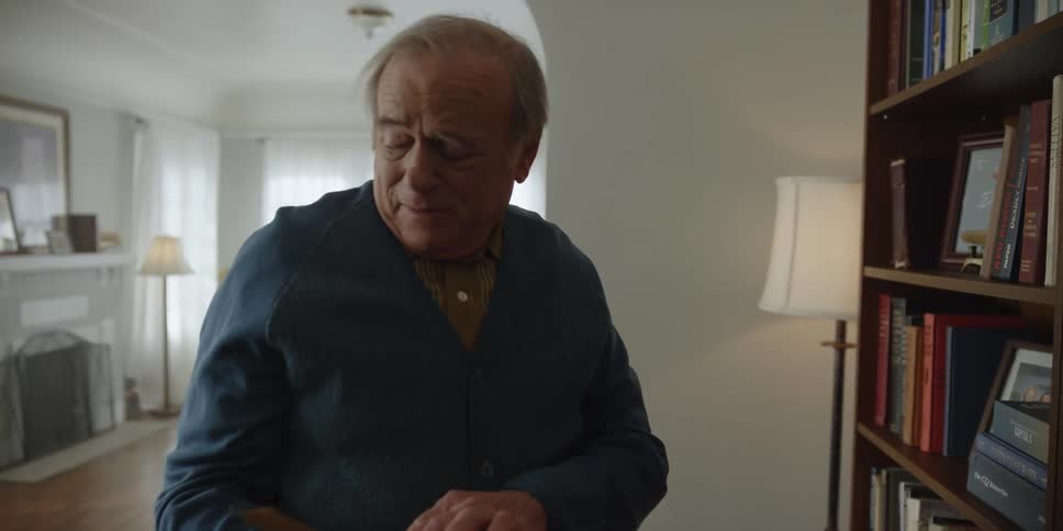 An elderly man at home looks at a book from a bookshelf as his wife sits in the background holding a piece of paper