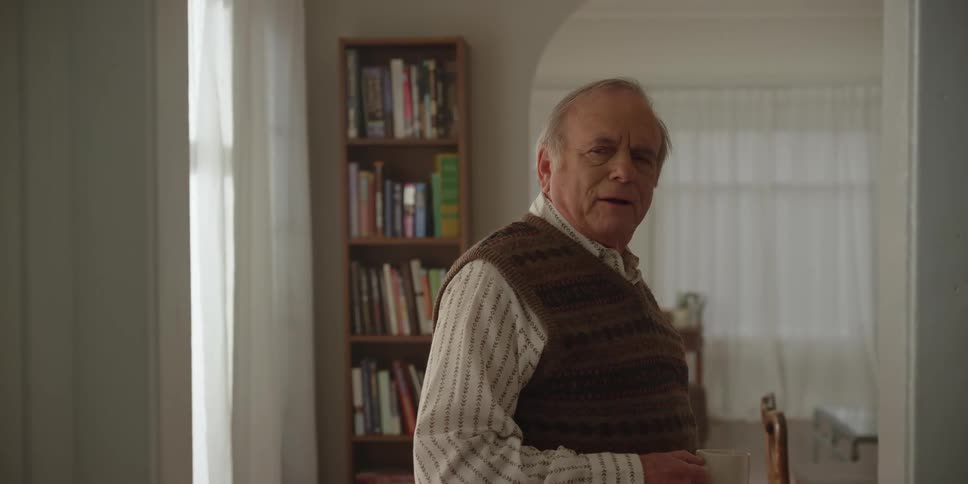 Elderly man talks to someone off screen before walking out of room while drinking from a cup