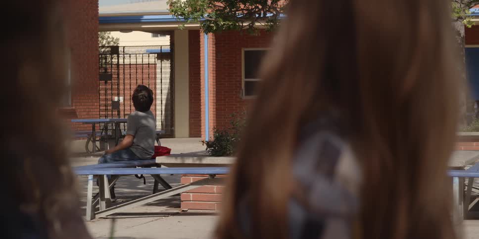 A young girl looks across a school yard at a boy sitting on a bench as another child runs past