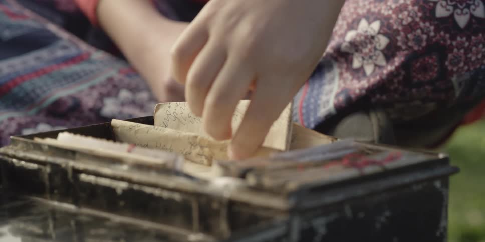 One person picks up old letters from ornate old box as another female sits cross legged on ground next to it