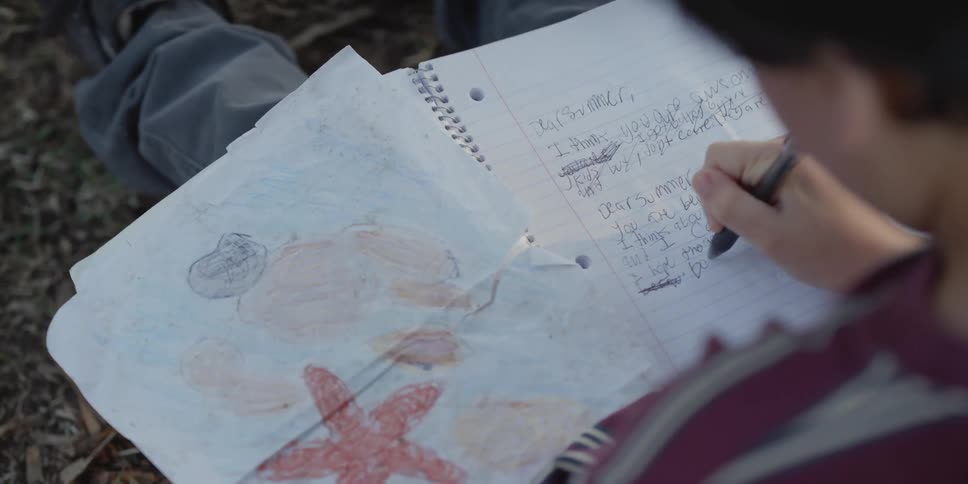 A young boy sits outside and writes a note to a girl