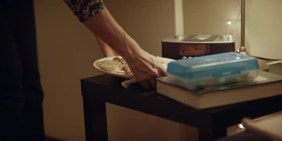 A person puts a plate of food on the nightstand then sits