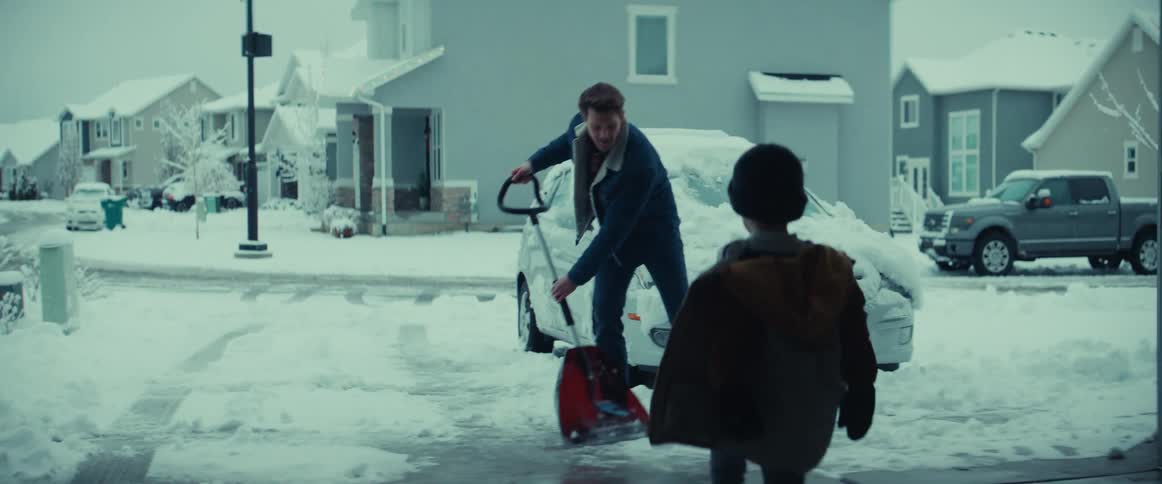 A boy walks out of a garage to approach a man shoveling snow from the driveway