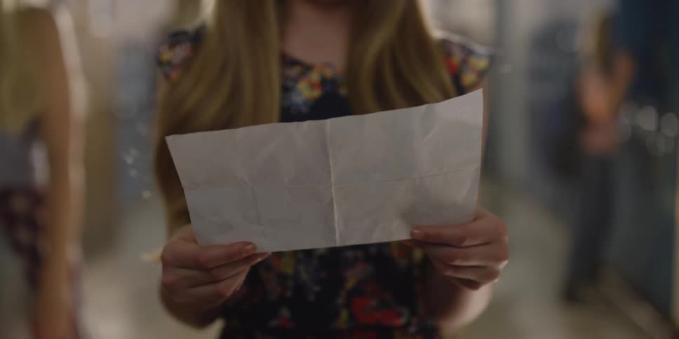A teenage girl is standing in a school hallway looking at a sheet of paper she is holding in her hands as people walk by her